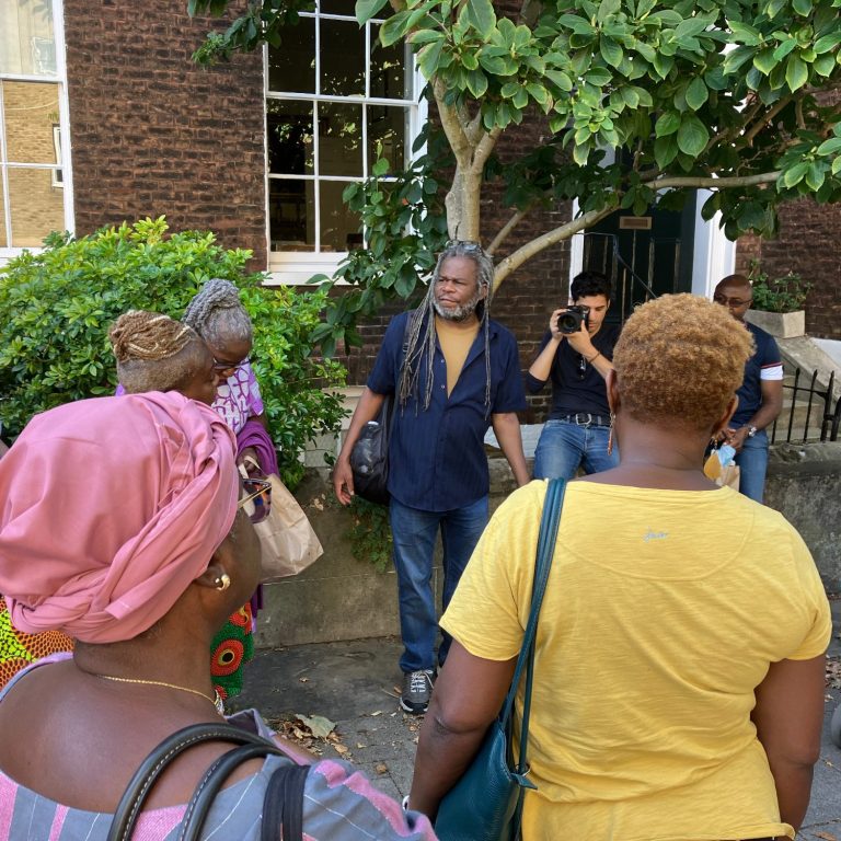 Historian S.I. Martin leading walking tour on Krios in Clapham, in collaboration with Iyamide Thomas (London Unseen, 2022) A group of people engaged in conversation on a tree-lined London street.